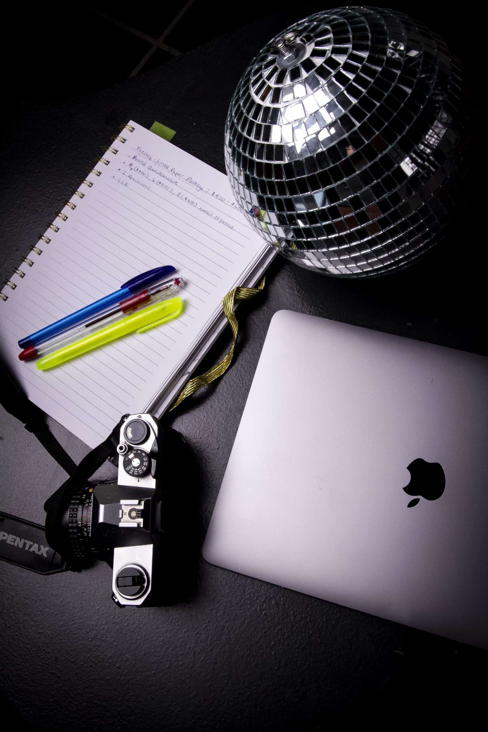 A laptop, notebook, pens, camera, and disco ball on a table